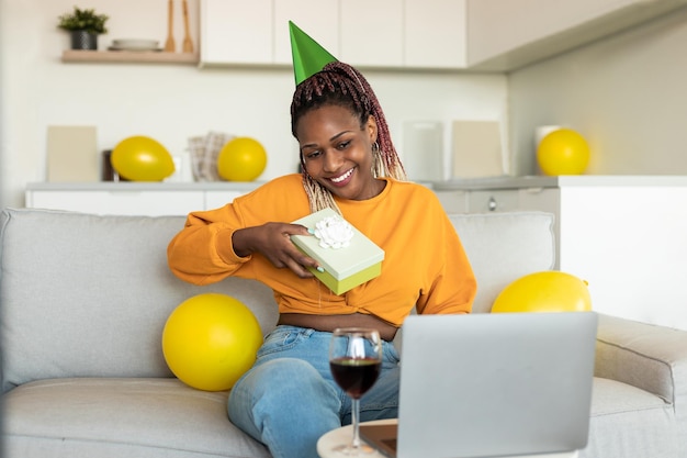 Feliz senhora afro-americana olhando para laptop recebendo presente de amigos comemorando aniversário via videochamada em casa