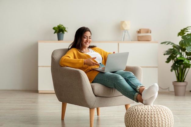 Feliz senhora árabe sentada na poltrona com laptop estudando remotamente ou videoconferência do espaço de cópia em casa