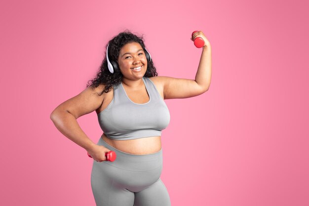 Foto feliz senhora gordinha afro-americana em fones de ouvido sem fio esportivos fazendo exercícios com halteres