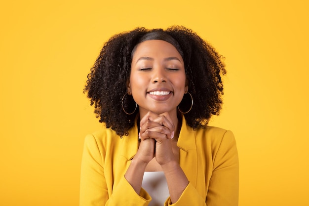 Feliz senhora negra sorridente fazendo gesto de oração fazendo um desejo pedindo milagre em pé no amarelo