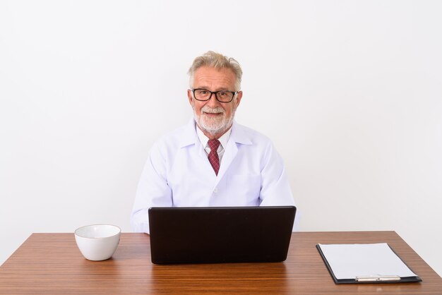 feliz sênior barbudo médico sorrindo e sentado na mesa de madeira com o laptop em branco