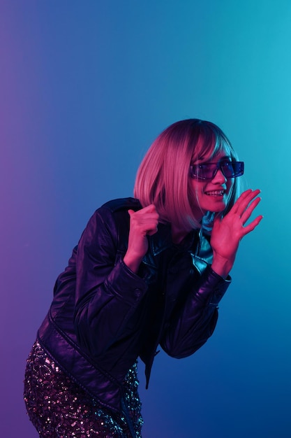 Feliz sexy hermosa mujer rubia en chaqueta de cuero vestido brillante gafas de sol de moda apuntar el pulgar hacia atrás dice chismes mirar a un lado posando aislado en color rosa azul fondo de estudio de luz Concepto de neón