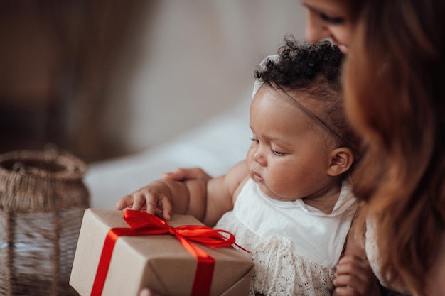 Feliz sincera madre auténtica e hija de raza mixta con caja de regalo en un acogedor apartamento