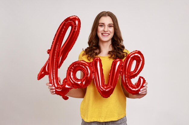 Feliz y sonriente adolescente romántica con camiseta informal amarilla sosteniendo letras de globos de aluminio, mirando a la cámara, expresando amor y gentileza. Disparo de estudio interior aislado sobre fondo gris.