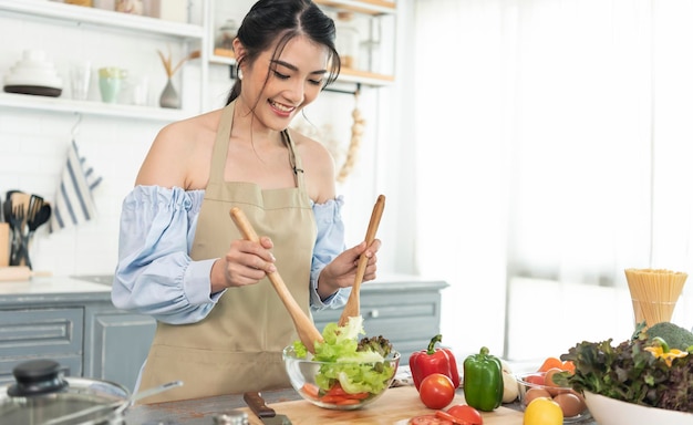 Feliz sonriente ama de casa asiática en delantal cocinando ensalada en la cocina mujer concepto de cocina saludable