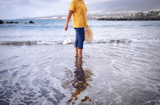 Feliz sonriente anciana caminando por la playa con sombrero y zapatos en las manos Señora madura descalza en el agua toda vestida mirando a la cámara Naturaleza escénica y horizonte en el fondo