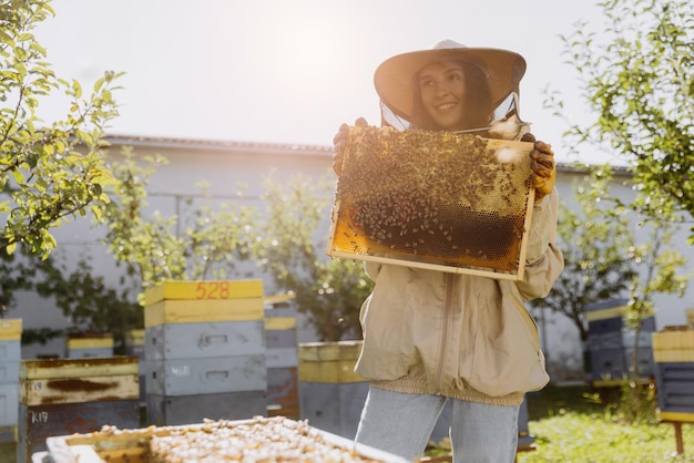 Feliz y sonriente apicultora en traje de protección sosteniendo el marco de la abeja con las abejas en el apiario