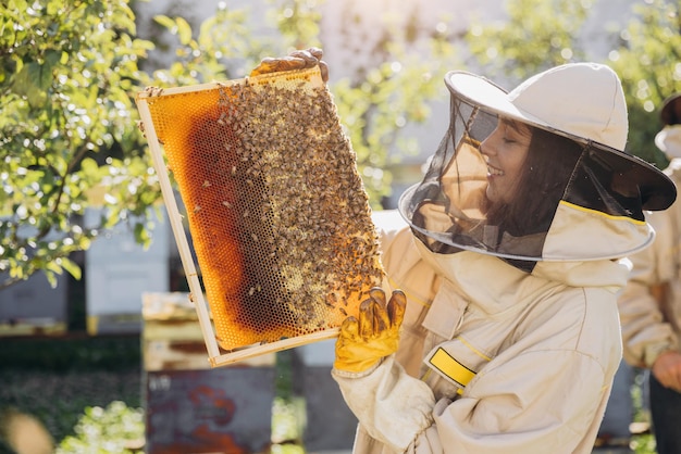 Foto feliz y sonriente apicultora en uniforme de pie en el apiario y sosteniendo el marco de la abeja