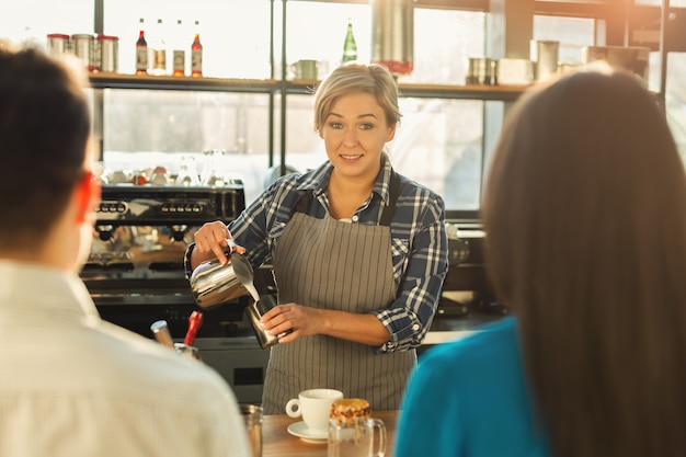 Feliz sonriente barista haciendo café y hablando con sus invitados en la barra del bar. Bartender femenino experimentado que da la bienvenida a la pareja. Pequeñas empresas, personas de ocupación y concepto de servicio, espacio de copia