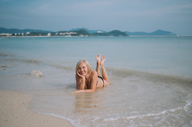 Feliz sonriente emocionado turista senior anciana jugando en el agua y nadando en las grandes olas en la playa del mar océano. Viajando por Asia, concepto de estilo de vida activo.