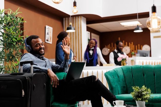 Feliz y sonriente empresario afroamericano sentado en el vestíbulo del hotel con equipaje mirando el portátil agitando la mano durante una videollamada mientras espera el check-in. asistir a una conferencia telefónica cuando viaja