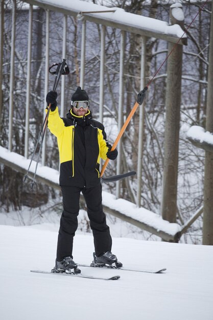 Feliz sonriente esquiador masculino usando t bar ski drag lift y saludando agitando sus bastones de esquí