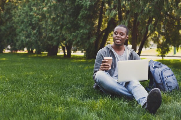 Feliz sonriente estudiante afroamericano sentado al aire libre en el césped con el portátil y tomar café, escribir, navegar por internet. Tecnología, comunicación, concepto de educación, espacio de copia