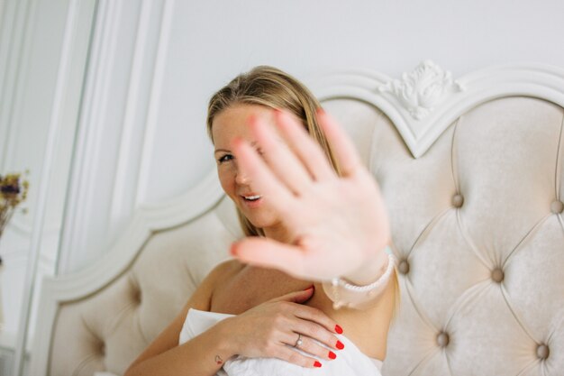 Feliz sonriente hermosa mujer joven de cabello largo tirando de la mano hacia la cámara sentado en la cama, enfoque selectivo en la mano