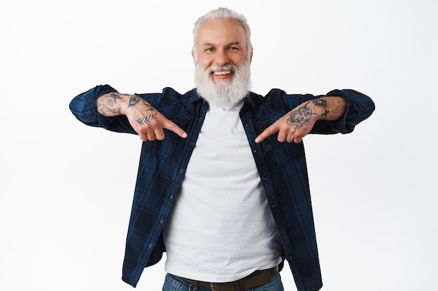 Foto feliz sonriente hombre mayor con barba y tatuajes, señalando con el dedo en el centro, mostrando el banner de su logotipo, mostrando el lugar para la publicidad, de pie sobre una pared blanca