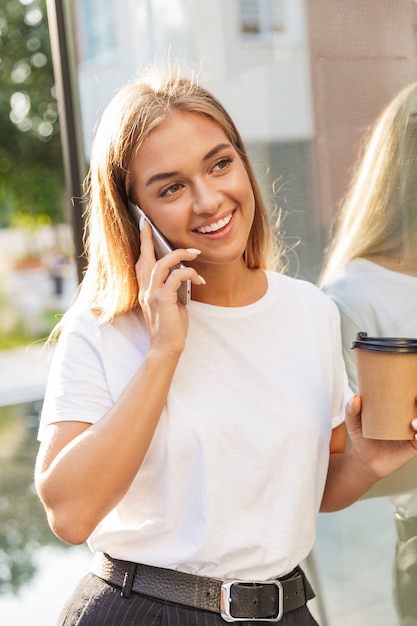 Feliz sonriente joven empresaria positiva con una taza de café posando al aire libre cerca del centro de negocios hablando por teléfono móvil.