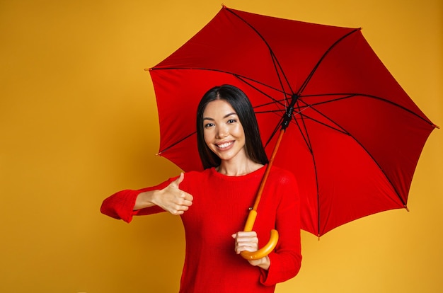 Feliz sonriente joven hermosa mujer asiática en ropa casual se para con paraguas rojo y mira a la cámara sobre fondo amarillo