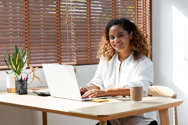 Feliz sonriente joven mujer de negocios afroamericana mirando la cámara mientras trabaja desde casa en una computadora portátil en la sala de estar