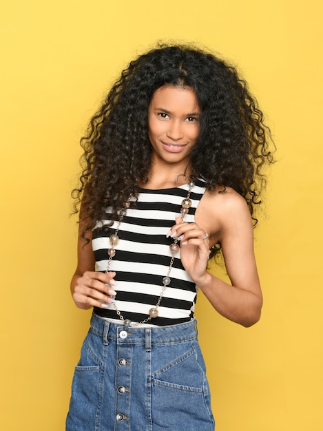 Foto feliz sonriente joven negra en falda de mezclilla y top de rayas con collar posando sobre un fondo amarillo mirando a la cámara con una linda sonrisa