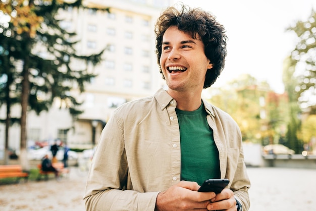 Feliz sonriente joven de pie al aire libre tiene una expresión alegre durante el juego en el teléfono móvil Joven hombre guapo con el pelo rizado navegando en línea en su teléfono inteligente en la calle de la ciudad