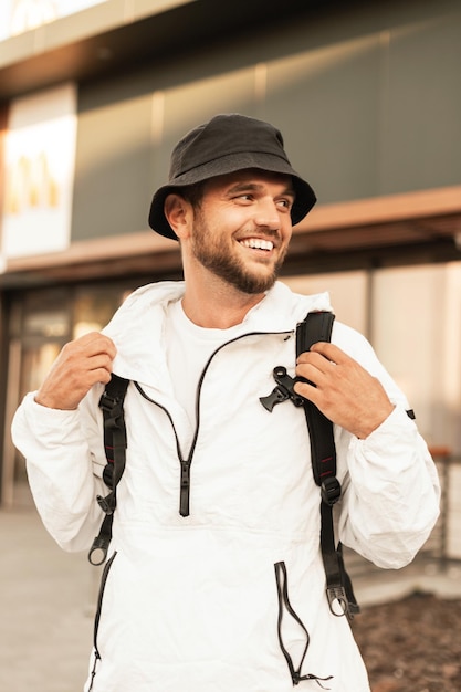 Feliz sonriente joven con sombrero de cubo de moda en ropa blanca con mochila camina y viaja por la ciudad