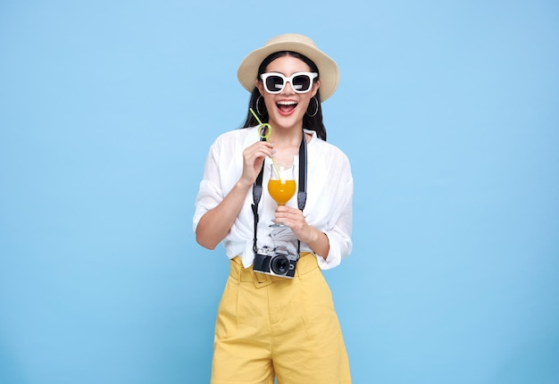 Feliz sonriente joven turista asiática con sombrero de verano de pie con cámara y bebiendo jugo de naranja