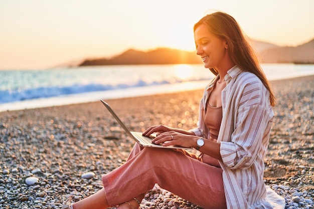 Feliz sonriente, linda, alegre, satisfecha, niña, independiente, trabajo remoto, en línea, en la orilla del mar, al atardecer, trabajo, por todas partes