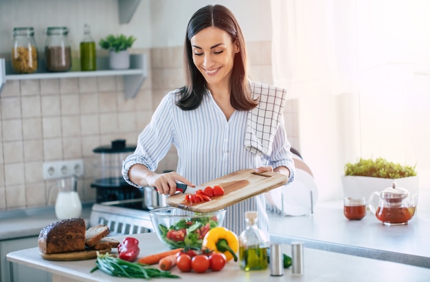 Feliz sonriente linda mujer está preparando una ensalada vegana saludable fresca con muchas verduras en la cocina de casa y está probando una nueva receta