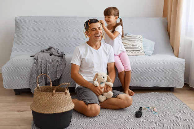 Feliz sonriente morena hombre vestido con estilo casual sentado en el piso, pasando tiempo juntos, niña haciendo peinado para el padre, aferrándose a mechones de colores en la cabeza de papá.