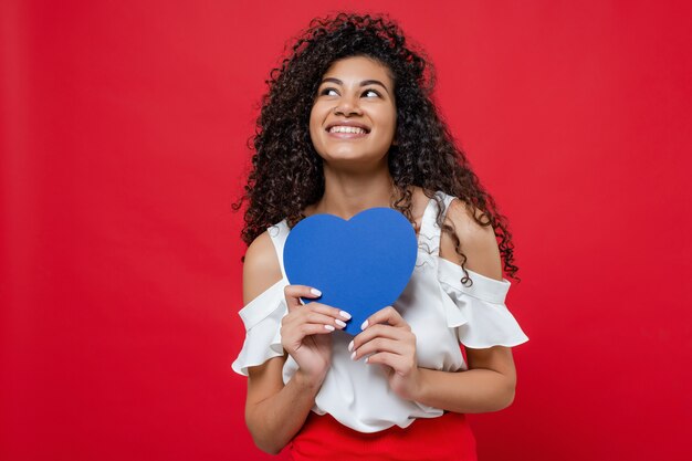 Feliz sonriente mujer africana con tarjeta de San Valentín en forma de corazón azul aislado en rojo