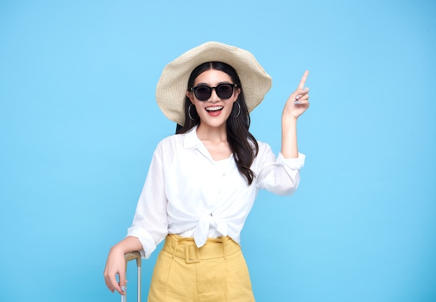 Feliz sonriente mujer asiática vestida con ropa de verano y con sombrero con equipaje disfrutando de sus vacaciones de verano y señalando con el dedo un espacio de copia en la pared azul brillante.