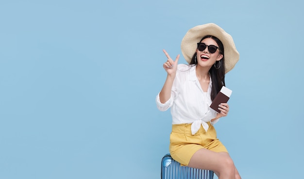 Feliz sonriente mujer asiática vestida con ropa de verano y con sombrero con equipaje disfrutando de sus vacaciones de verano y señalando con el dedo un espacio de copia sobre fondo azul brillante.