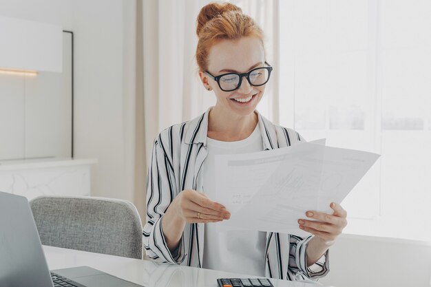 Feliz sonriente mujer caucásica leyendo buenas noticias en documentos financieros mientras gestiona el presupuesto familiar