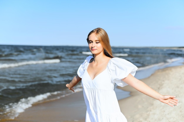 Feliz y sonriente mujer hermosa está en la playa del océano con un vestido blanco de verano, con los brazos abiertos.