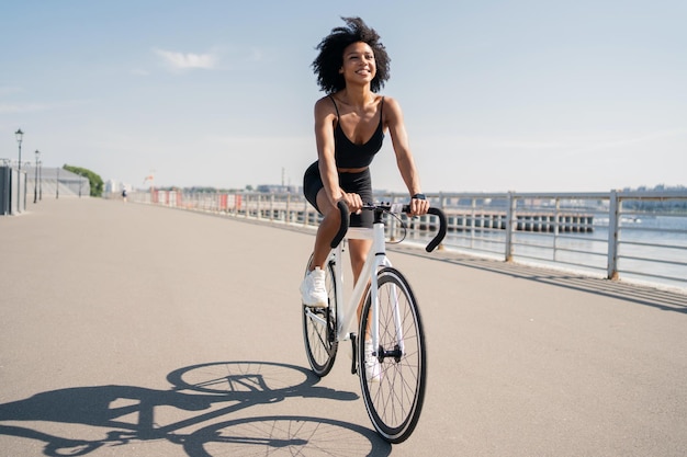 Feliz sonriente mujer joven de pelo rizado actividad contenta bicicleta de carretera montando en un traje negro deportivo