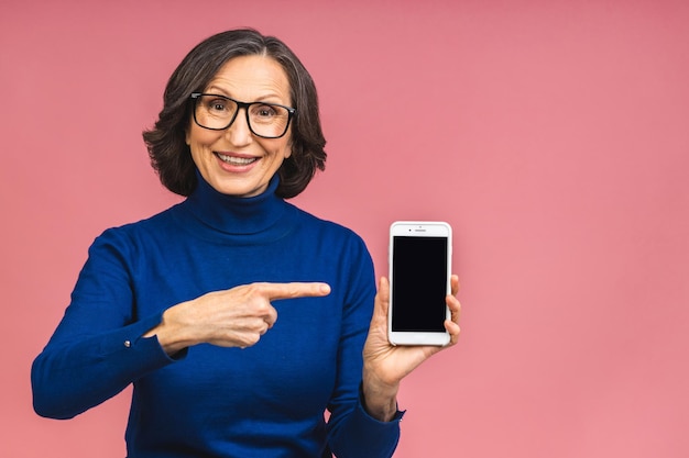 Feliz sonriente mujer madura senior feliz en casual mostrando la pantalla del teléfono inteligente en blanco mientras mira a la cámara aislada sobre fondo rosa. Usando el teléfono.