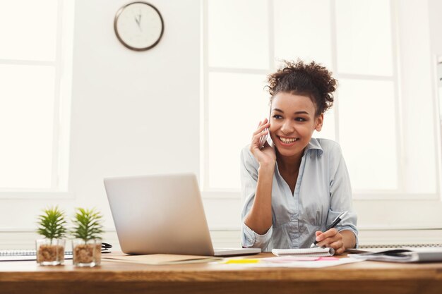 Feliz sonriente mujer de negocios afroamericana en el trabajo hablando por teléfono y tomando notas, sentada en su lugar de trabajo en la oficina, espacio de copia