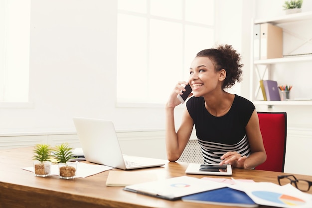 Feliz sonriente mujer de negocios en el trabajo hablando por teléfono, sentada en su lugar de trabajo en la oficina y usando tableta digital, espacio de copia