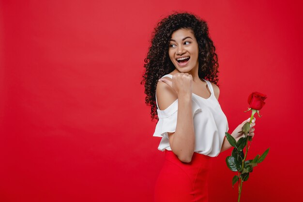 Foto feliz sonriente mujer negra con rosa roja en la mano aislada sobre rojo