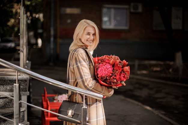 Feliz y sonriente mujer rubia en abrigo a cuadros con un gran ramo de flores rojas