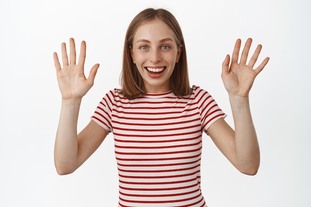 Feliz sonriente mujer rubia agitando las manos, decir adiós o hola, saludo o despedida, luciendo amable y alegre, de pie con una camiseta a rayas sobre una pared blanca