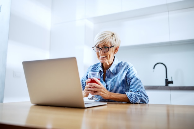 Feliz sonriente mujer senior rubia sentada en la mesa del comedor, bebiendo vino tinto y con videollamada en la computadora portátil