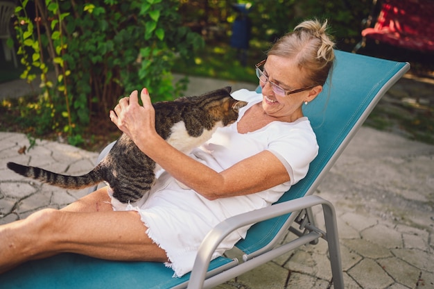 Feliz sonriente mujer senior en vasos de relax en el jardín de verano al aire libre, abrazando gato atigrado