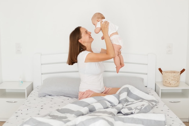 Feliz sonriente mujer vestida con camiseta blanca de estilo casual sosteniendo a la niña en las manos mientras está sentada en la cama en la habitación luminosa, mamá pasa tiempo con su bebé recién nacido.