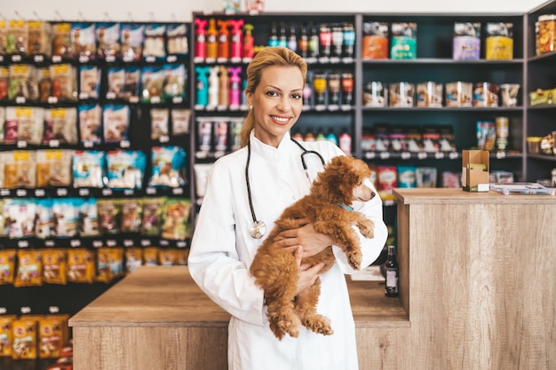 Foto feliz y sonriente mujer veterinaria de mediana edad de pie en la tienda de mascotas y sosteniendo un lindo caniche rojo en miniatura mientras mira a la cámara.