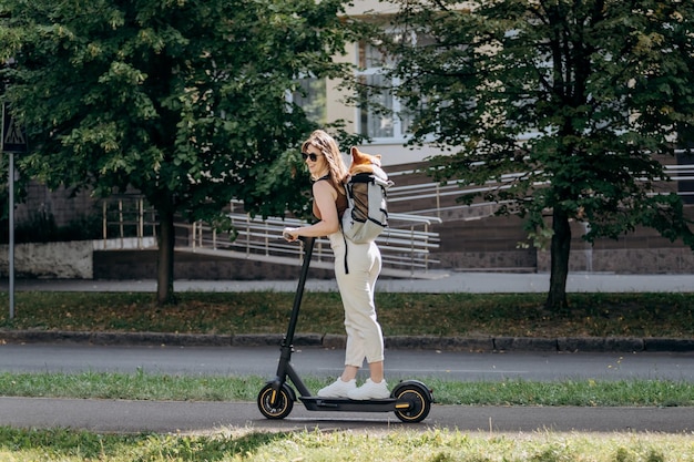 Feliz sonriente mujer viajera está montando su electro scooter en el parque de la ciudad con el perro Welsh Corgi Pembroke en una mochila especial