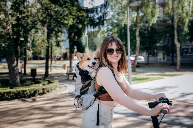 Feliz sonriente mujer viajera está montando su electro scooter en el parque de la ciudad con el perro Welsh Corgi Pembroke en una mochila especial