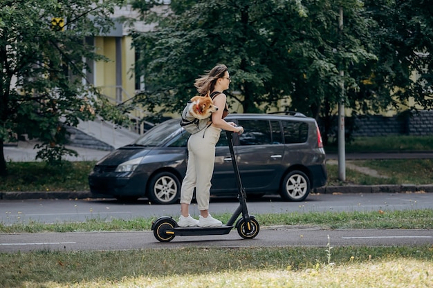 Feliz sonriente mujer viajera está montando su electro scooter en el parque de la ciudad con el perro Welsh Corgi Pembroke en una mochila especial