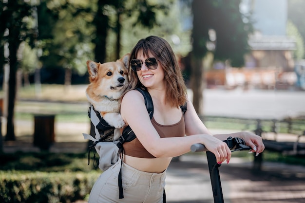 Feliz sonriente mujer viajera está montando su electro scooter en el parque de la ciudad con el perro Welsh Corgi Pembroke en una mochila especial