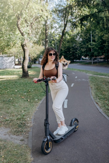 Feliz sonriente mujer viajera está montando su electro scooter en el parque de la ciudad con el perro Welsh Corgi Pembroke en una mochila especial